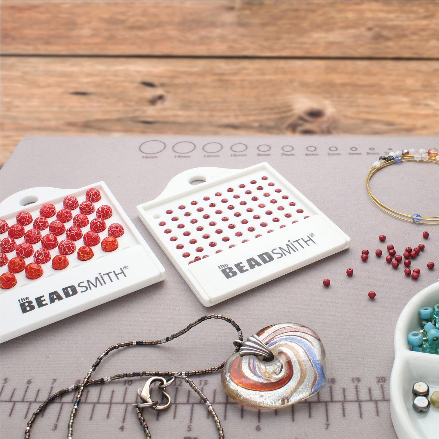 Jewelry making supplies on a mat: red beads, The Beadsmith bead trays, bead strands, and a pendant.