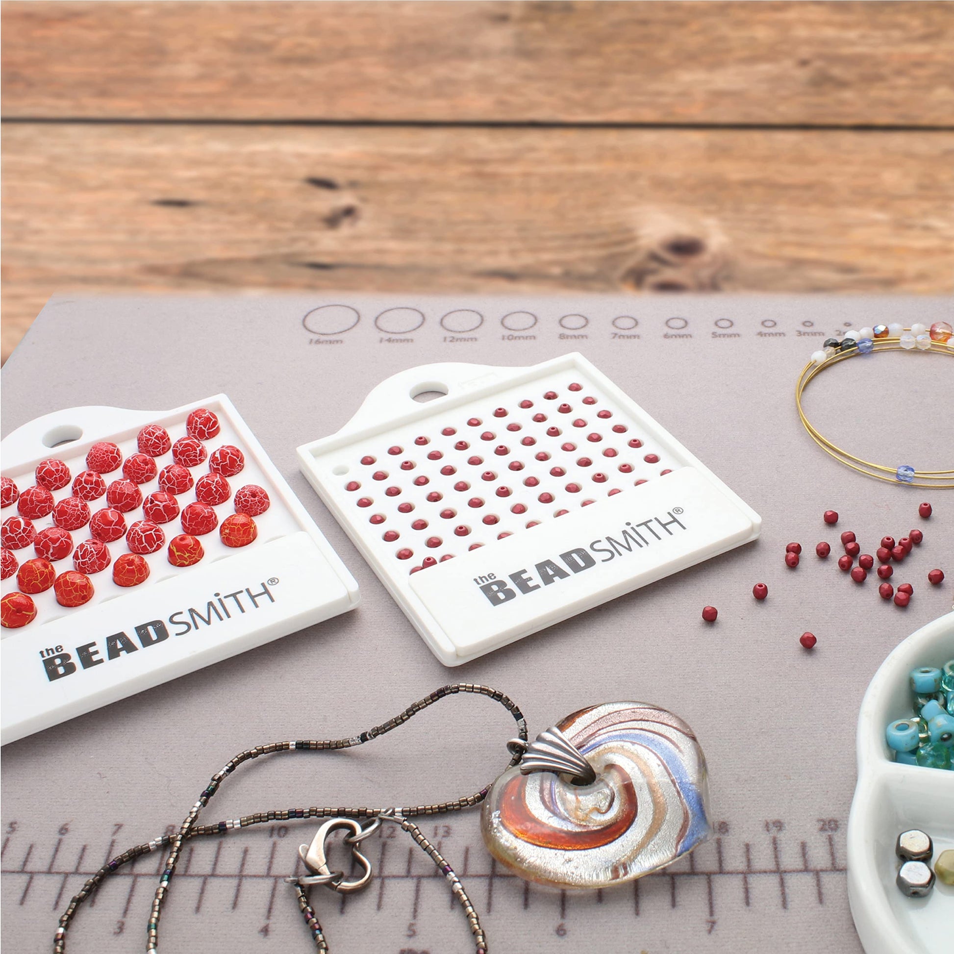 Jewelry making supplies on a mat: red beads, The Beadsmith bead trays, bead strands, and a pendant.